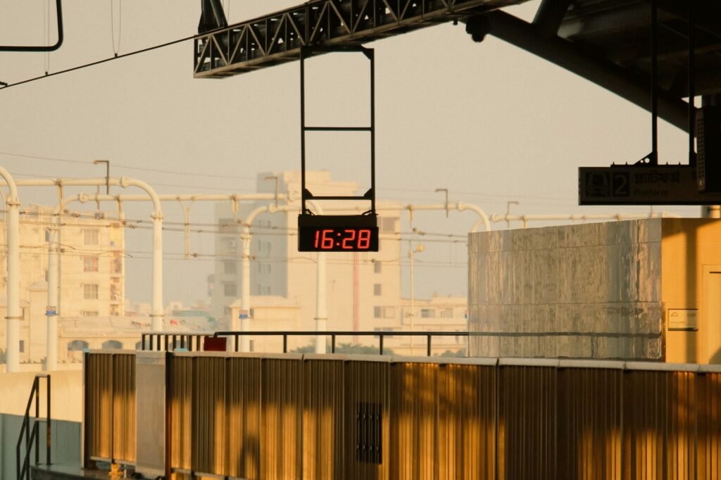Digital clock displaying 16:28 at a railway station during golden hour, illuminated by warm sunset light.