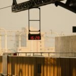Digital clock displaying 16:28 at a railway station during golden hour, illuminated by warm sunset light.