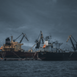 Cargo ships at a bustling port under cloudy skies, cranes ready for loading.