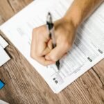 Hand writing on a W-9 tax form using a ballpoint pen on wooden desk.