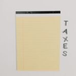 Close-up of a yellow legal pad and the word 'TAXES' on a white surface, ideal for finance themes.