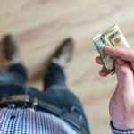 A person wearing denim and a checked shirt holds folded dollar bills in their hand indoors.