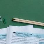 U.S. tax forms with pencils and paperclips on green surface in a flat lay setup.