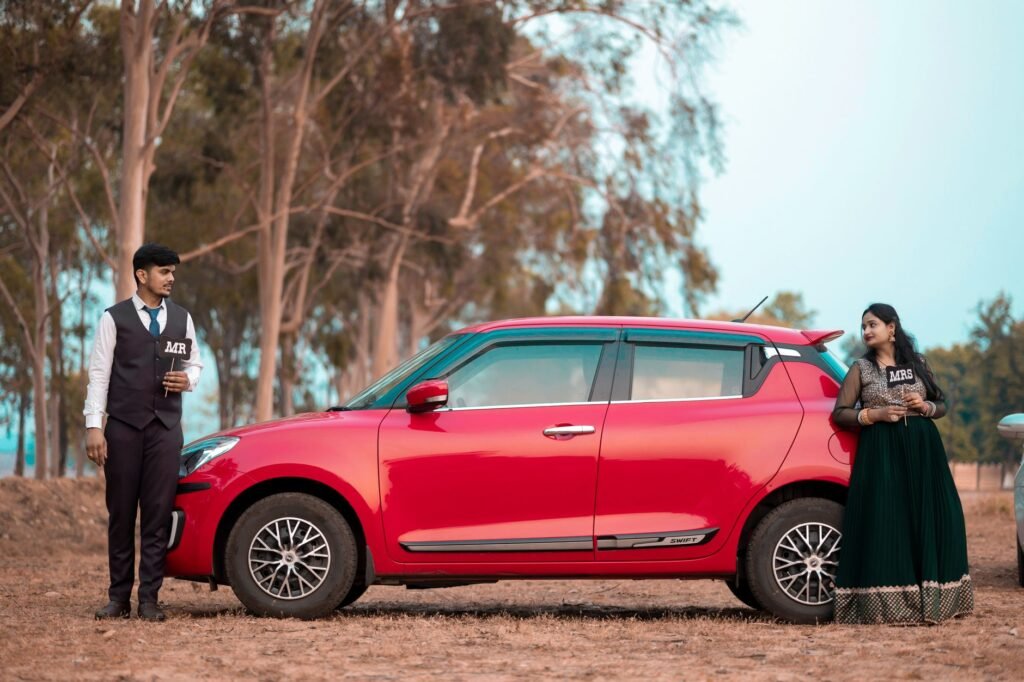 Stylish Indian couple in traditional attire posing by a red car in a rural setting.
