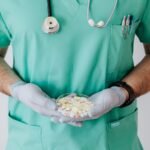 Unrecognizable male doctor wearing medical uniform and latex gloves holding Petri dish filled with assorted drugs while standing near white wall in hospital