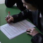 A woman in a suit uses a red pen to edit a printed document on a green table with a laptop.