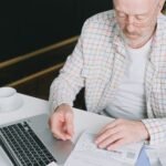 Elderly man with eyeglasses reviewing documents at a laptop. Indoor setting with natural light.