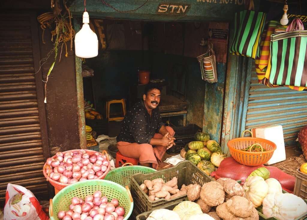 A market vendor smiling amidst a vibrant display of fresh vegetables and fruits.