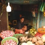 A market vendor smiling amidst a vibrant display of fresh vegetables and fruits.