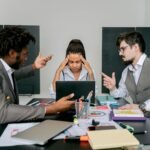 Two men argue while a woman looks frustrated at a laptop in an office environment.
