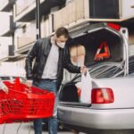 Young father and little son in casual clothes and protective masks moving shopping bags from supermarket trolley to trunk modern building parking