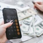 Close-up of hands using smartphone calculator next to dollar bills on table. Financial planning concept.