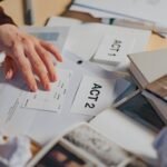 A cluttered desk with scripts, notes, and a person's hand organizing screenplay drafts.