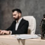 A businessman working on a laptop in an office with a Lady Justice statue in focus on the desk.