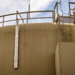 A low angle view of a large industrial water tank with a measuring scale and ladder under a clear sky.
