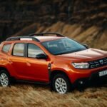 A second-generation red Dacia Duster SUV parked on a rural roadside, showcasing its subcompact crossover design.
