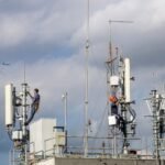Two workers repairing a cellular tower against a cloudy sky, with an airplane in the background.