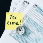 A stack of tax forms with a clock and yellow sticky note saying 'Tax time!' indicating urgency.
