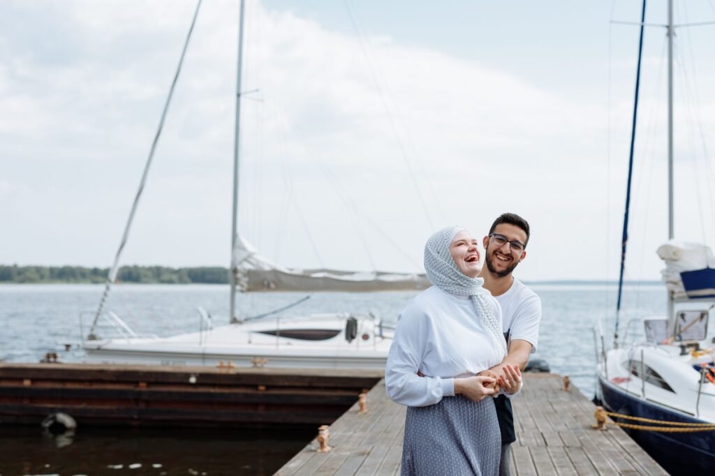 Joyful couple laughing together on a wooden dock by the marina, surrounded by boats and clear skies.