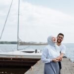 Joyful couple laughing together on a wooden dock by the marina, surrounded by boats and clear skies.