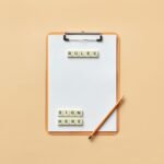 Flat lay of clipboard with letter tiles spelling 'rules' and 'sign here', accompanied by pencil on beige background.