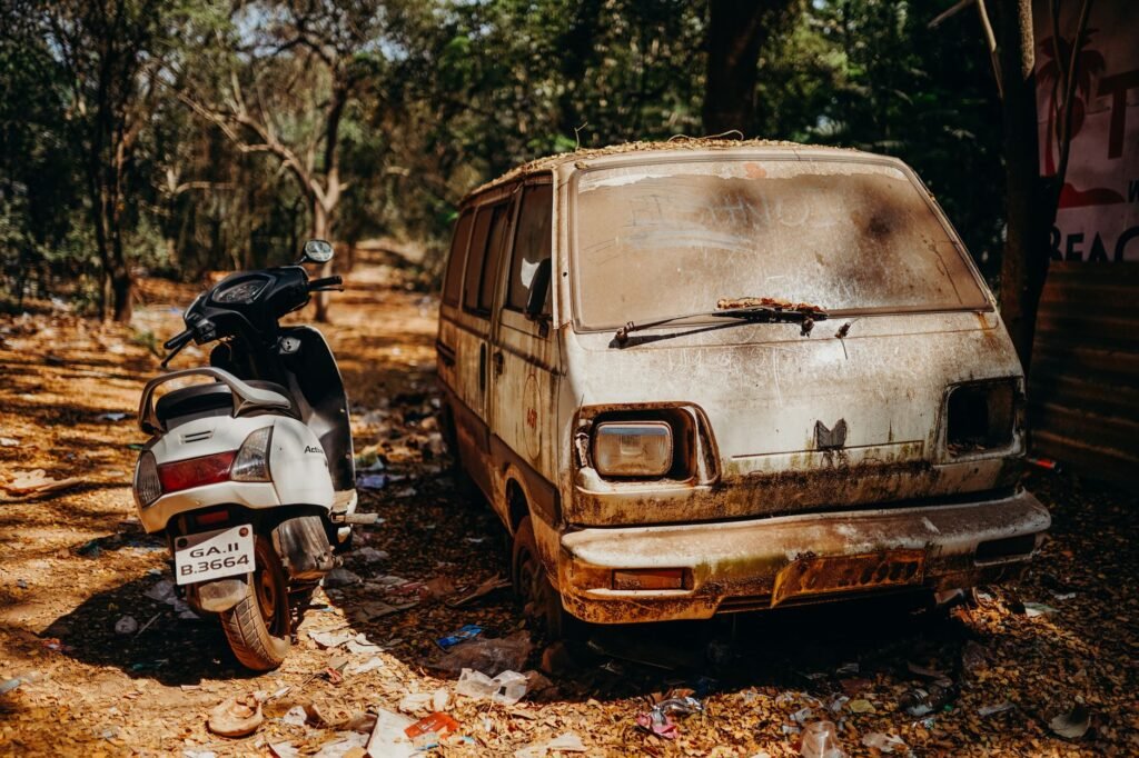 Abandoned rusty van and scooter under trees on a sunny day, highlighting decay and nature.