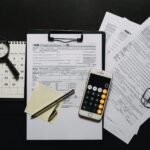 Top view of tax documents, calculator, magnifying glass, and calendar on a black surface.