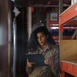 Woman in warehouse taking inventory with clipboard, focused and pensive.