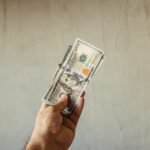A hand holding a stack of US dollar banknotes against a neutral backdrop, symbolizing wealth.