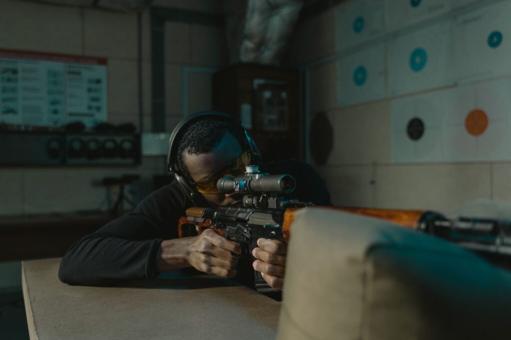 A determined man wearing ear and eye protection aims a rifle at a target in an indoor shooting range.