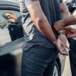 A man being handcuffed by police near a car, with reflection visible.