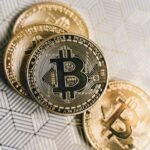 A close-up image of gold Bitcoin cryptocurrency coins on a geometric patterned surface.