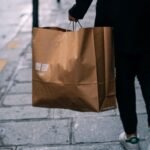 A person walking on the street carrying a brown paper shopping bag, symbolizing urban shopping.