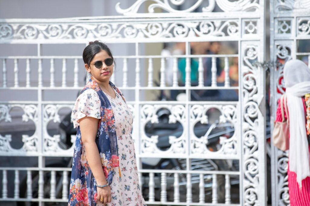 Modern woman in sunglasses poses by a decorative white metal gate on a sunny day.