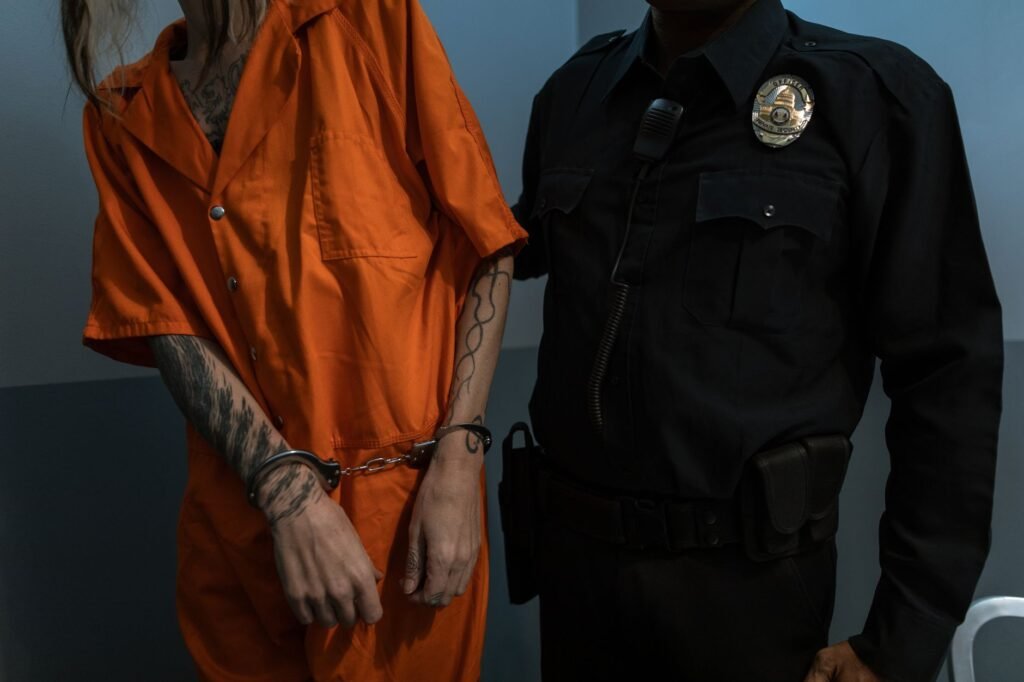 Tattooed prisoner handcuffed in orange jumpsuit beside uniformed officer, highlighting law enforcement.