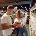 From above of young woman in casual wear showing canned food for man while standing with grocery trolley near shelves with products in modern supermarket