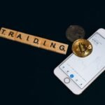 Cryptocurrency trading concept featuring Bitcoin, Scrabble tiles, and a smartphone on a black surface.