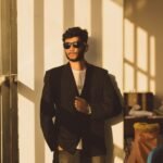 A fashionable man wearing sunglasses and a suit leans confidently against a sunlit wall.